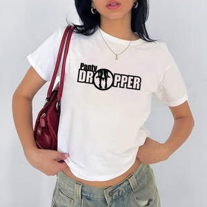 Könnte beinhalten: Weißes T-Shirt mit dem schwarzen Text "Panty Dropper" und einer kreisförmigen Grafik. Das kurzärmelige Shirt scheint aus einem weichen Material zu sein. Eine rote Handtasche ist im Bild zu sehen.