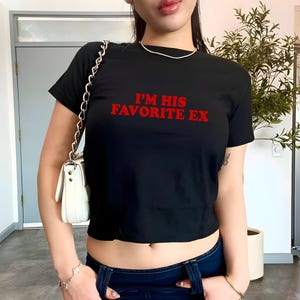 Könnte beinhalten: Schwarzes Crop-Top mit dem roten Text "I'M HIS FAVORITE EX". Das Shirt hat kurze Ärmel und einen Rundhalsausschnitt. Das Model trägt eine blaue Jeans und eine weiße Handtasche mit einer silbernen Kette.