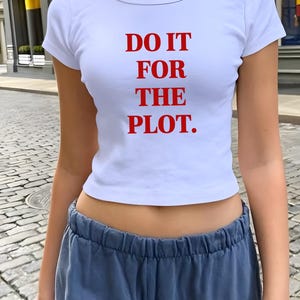 Puede incluir: Top corto blanco con el texto rojo "DO IT FOR THE PLOT." La camiseta tiene cuello redondo y mangas cortas. La modelo lleva un pantalón de chándal azul. El fondo es una calle adoquinada.