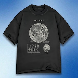 May include: Black t-shirt with a detailed white illustration of the moon and its phases. The design includes the words "THE MOON" and translations in French, German, and Spanish. The t-shirt has a round neckline and short sleeves.
