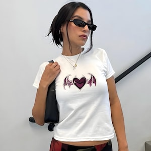 May include: White t-shirt featuring a dark purple heart with stylized purple wings. The graphic is centered on the front of the short-sleeved shirt. The shirt is made of a soft, lightweight material.