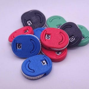 May include: A collection of colorful, numbered weight plates in red, blue, green, and black. Each plate has a white number and a curved groove. The plates are stacked together on a white surface.