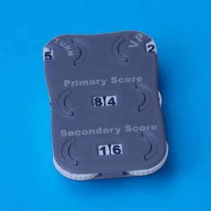 May include: A gray plastic game score tracker with white trim, set against a blue background. The tracker displays "Primary Score" with a score of 84 and "Secondary Score" with a score of 16. It also has "TURN" and "VP" indicators.
