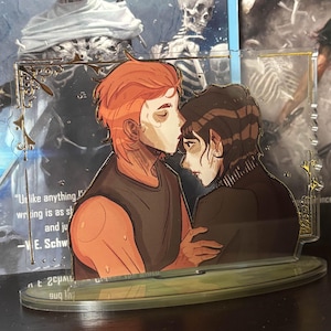 May include: A clear acrylic stand featuring an illustration of two figures embracing, with one figure kissing the other's forehead. The stand has a decorative gold border. The background includes a book with text and a skeletal figure.