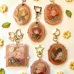 Może przedstawiać: Kolekcja akrylowych breloków do kluczy z ilustracjami postaci w stylu anime w różnych pozach. Każdy brelok ma złote zapięcie i dekoracyjną ramkę. Breloki otoczone są małymi żółtymi kwiatami i zielonymi liśćmi.