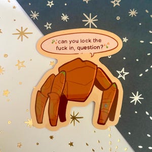 May include: A die-cut sticker featuring a stylized orange crab-like creature with segmented legs. Above the creature is a speech bubble containing the text, "can you lock the fuck in, question?" with small musical note icons on either side.