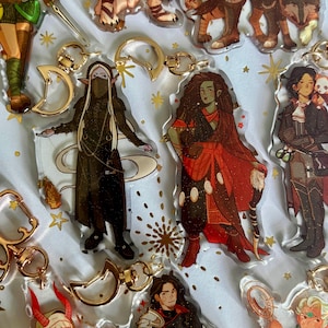 May include: A collection of clear acrylic keychains featuring various illustrated fantasy characters with gold-toned moon-shaped clasps. The characters include figures in dark robes, red garments, and armor, all accented with subtle glitter details.