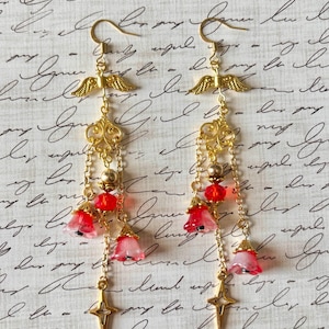 Peut inclure: Boucles d'oreilles pendantes dorées sur le thème céleste. Chaque boucle d'oreille présente un charme ailé, des filigranes ornés, des perles rouges et de délicats charmes de fleurs roses et rouges. Un pendentif étoile dorée est suspendu en bas.