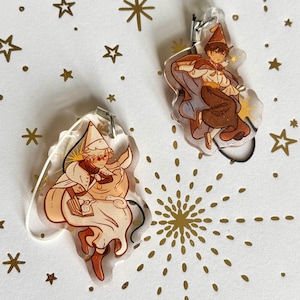 May include: Two clear acrylic charms featuring illustrated characters in pointed hats. One charm depicts a light-haired figure in a cream-colored robe holding a book, while the other shows a dark-haired figure in a brown outfit and cape.