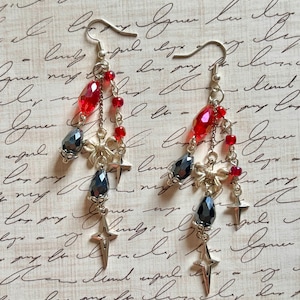 May include: Dangling earrings with silver hooks, featuring red and dark gray faceted beads, small red beads, and star-shaped charms. The earrings have a delicate, layered design with a vintage aesthetic.
