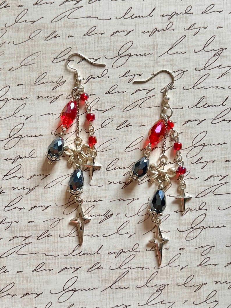 May include: Dangling earrings with silver hooks, featuring red and dark gray faceted beads, small red beads, and star-shaped charms. The earrings have a delicate, layered design with a vintage aesthetic.