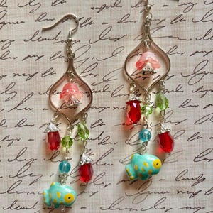 May include: A pair of dangle earrings with silver-tone metalwork. Each earring features a pink flower, red and green beads, and a turquoise fish charm with yellow spots. The earrings are on a patterned background.