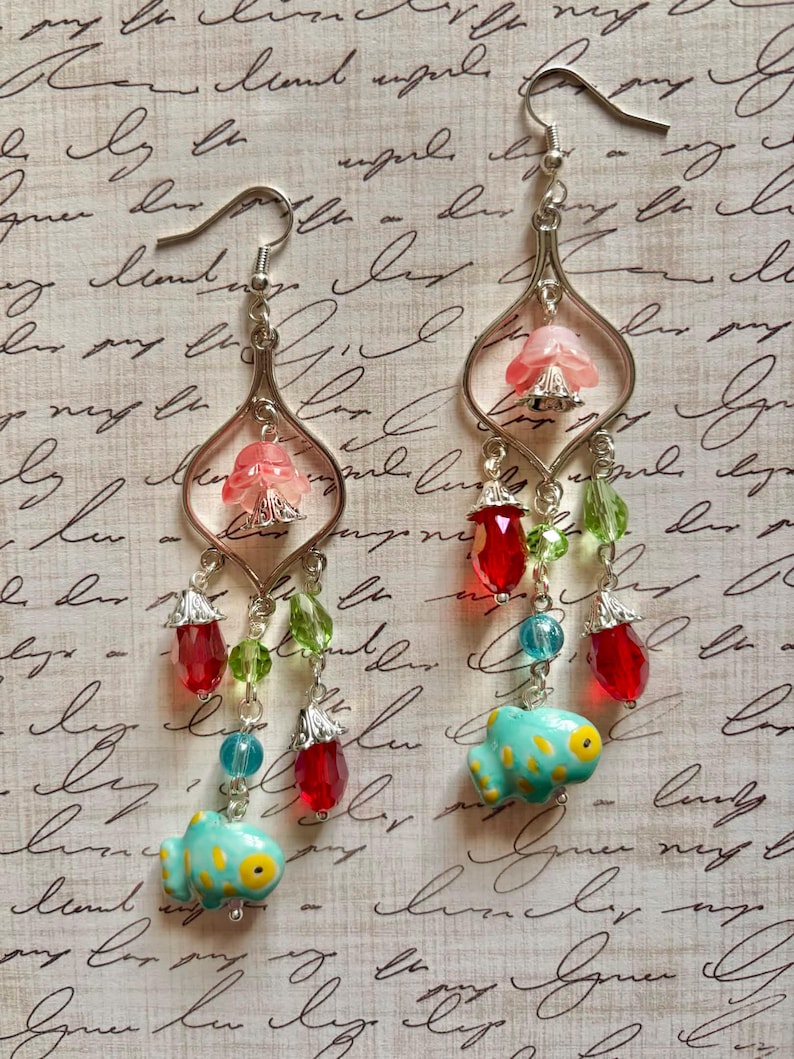 May include: A pair of dangle earrings with silver-tone metalwork. Each earring features a pink flower, red and green beads, and a turquoise fish charm with yellow spots. The earrings are on a patterned background.