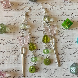 Peut inclure: Boucles d'oreilles pendantes au design fantaisiste. Chaque boucle d'oreille présente un charme d'aiguille argenté, un papillon rose, une feuille verte et des perles vertes. Les boucles d'oreilles sont agrémentées de boutons verts et de perles transparentes. Le fond comprend des dés.