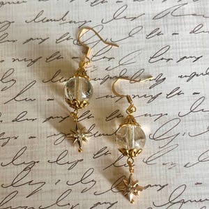 Pode incluir: Um par de brincos pendentes em tom dourado com uma conta esférica transparente embutida em tampas de filigrana dourada. Abaixo de cada conta, pende um pequeno pingente de estrela de oito pontas com minúsculos cristais transparentes.