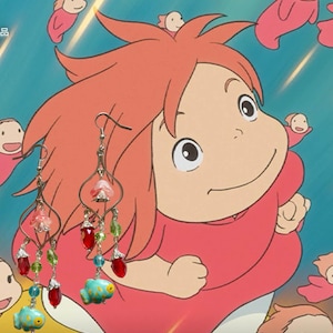 May include: A pair of dangle earrings featuring a whimsical design. The earrings have a silver-tone metal frame with pink, red, and green beads, and a turquoise fish charm. The background features a cartoon character.