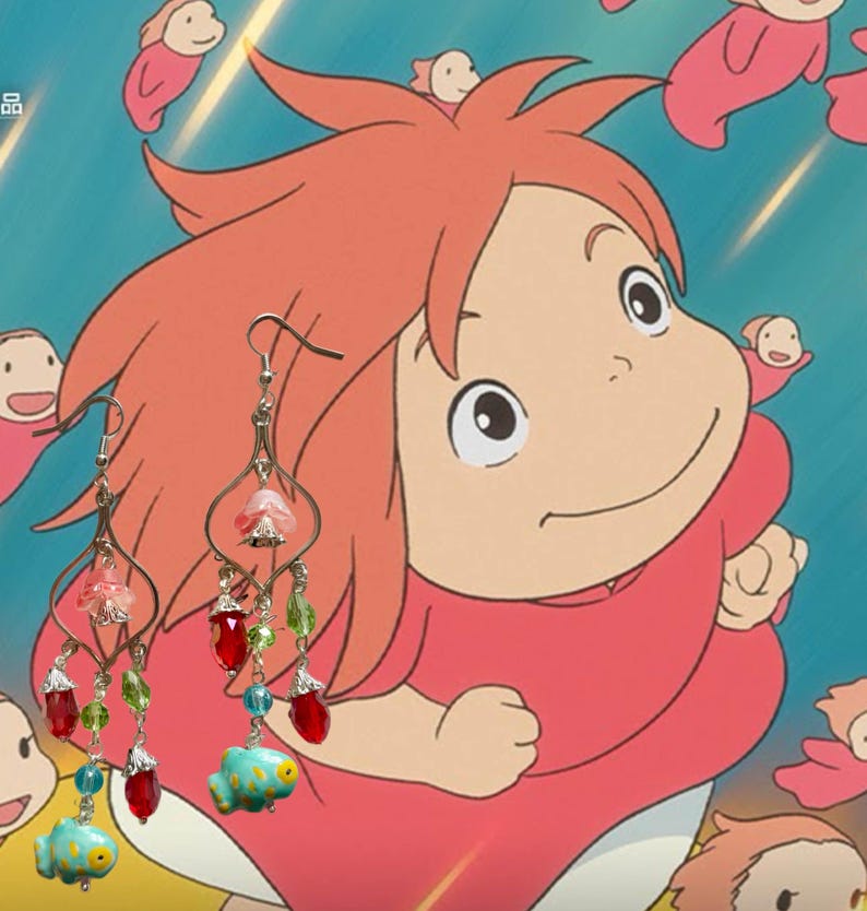 May include: A pair of dangle earrings featuring a whimsical design. The earrings have a silver-tone metal frame with pink, red, and green beads, and a turquoise fish charm. The background features a cartoon character.
