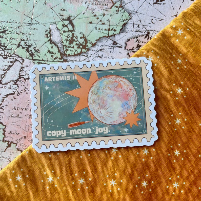 May include: A sticker with a postage stamp design featuring a colorful planet, orange starbursts, and the text "ARTEMIS II" and "copy moon joy." The sticker is on a yellow fabric with white star patterns, set against a map background.