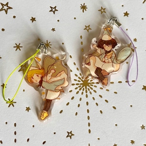 May include: Two clear acrylic charms featuring illustrated characters wearing party hats and capes. One charm has a bright yellow cord, and the other has a light purple cord. The characters are depicted in dynamic poses with brown and tan clothing.