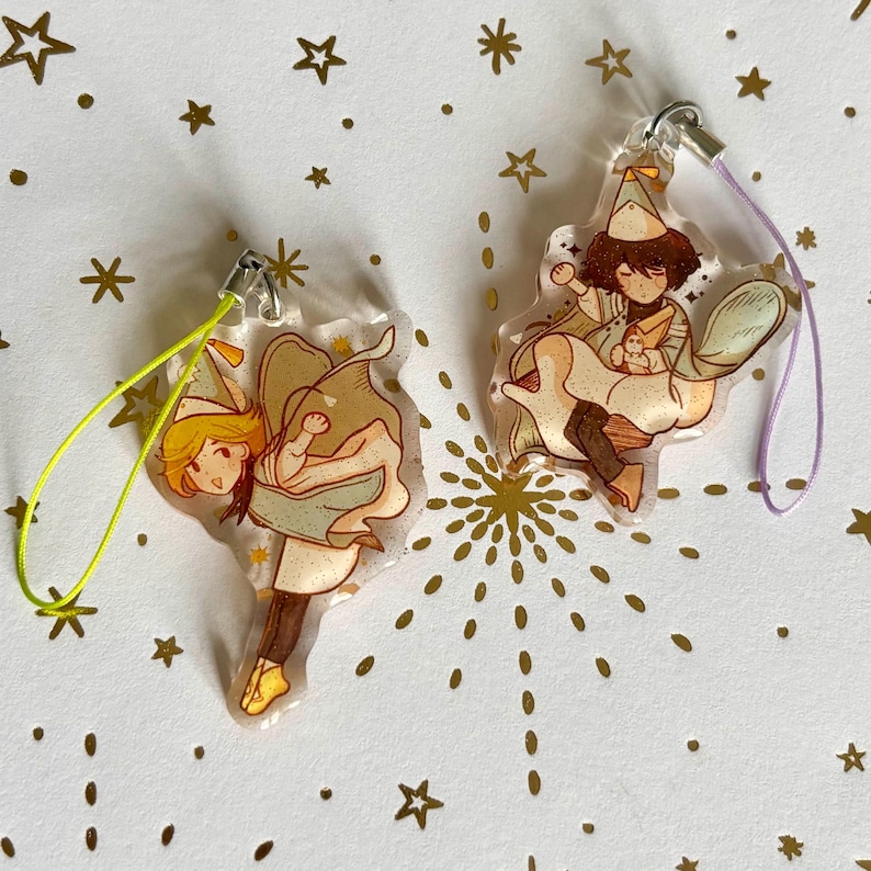 May include: Two clear acrylic charms featuring illustrated characters wearing party hats and capes. One charm has a bright yellow cord, and the other has a light purple cord. The characters are depicted in dynamic poses with brown and tan clothing.