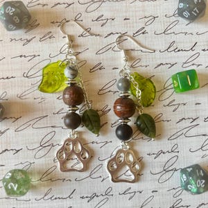 Peut inclure: Paire de boucles d'oreilles pendantes avec crochets argentés. Chaque boucle présente un charme en forme de patte, des perles en bois, des feuilles vertes et des perles grises et marron. Dés en arrière-plan.