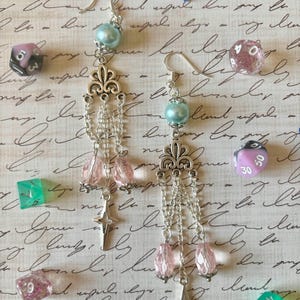 Peut inclure: Boucles d'oreilles argentées avec une perle bleu clair, un motif décoratif en argent, des perles de cristal roses et un charm en forme d'étoile argentée. Les boucles d'oreilles sont présentées avec divers dés colorés sur un fond manuscrit.