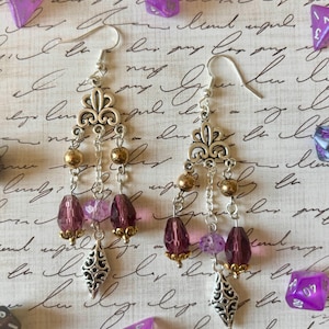 Peut inclure: Boucles d'oreilles pendantes argentées avec un motif de fleur de lys. Les boucles d'oreilles comportent des perles dorées, des perles violettes facettées et un charm décoratif argenté. Des dés violets sont dispersés autour des boucles d'oreilles.