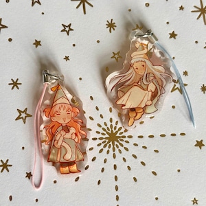 May include: Two clear acrylic charms featuring illustrated characters in pointed hats. The left charm has a pink cord and depicts a character with orange hair holding a small object. The right charm has a blue cord and shows a character with long hair.