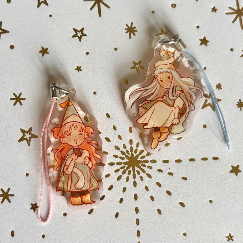 May include: Two clear acrylic charms featuring illustrated characters in pointed hats. The left charm has a pink cord and depicts a character with orange hair holding a small object. The right charm has a blue cord and shows a character with long hair.