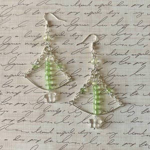 May include: A pair of handmade earrings featuring a silver-tone metal design with green and clear beads. The earrings have a geometric shape with a chain and hook closure. The background is a light beige color with script writing.