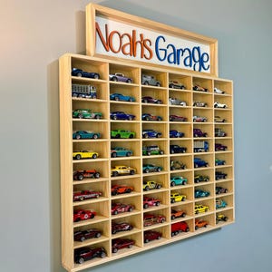 May include: Wooden display case with multiple compartments, filled with various toy cars. The case is mounted on a wall and features a sign that reads "Noah's Garage" in orange and blue lettering.