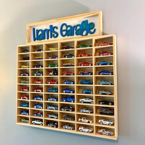 May include: A light-colored wooden wall-mounted display shelf with multiple rectangular compartments containing various miniature toy cars. Above the shelf, a wooden sign features the text "Liam's Garage" in blue and teal layered lettering.