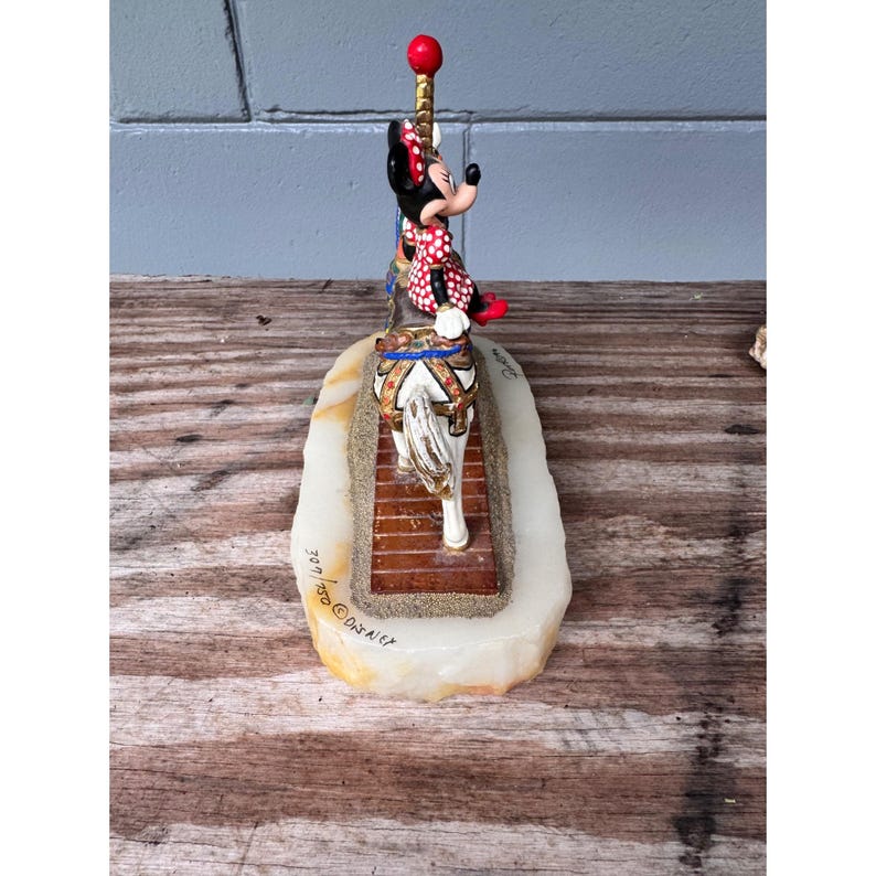 K&ouml;nnte beinhalten: Eine Minnie-Maus-Figur auf einem Karussellpferd, auf einem rechteckigen Sockel. Minnie tr&auml;gt ein rotes Kleid mit Tupfen und eine Schleife. Der Sockel ist ein hellfarbiger Stein mit sandiger Textur und einem h&ouml;lzernen Karussellboden. Die Figur hat eine rote Kugel oben.