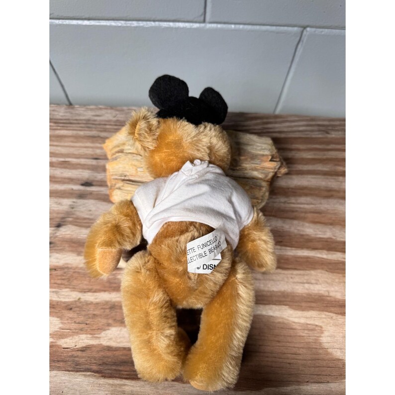 May include: A vintage teddy bear with a black bow and a white shirt. The bear has a tan fur and a tag that reads "Annette Funicello Collectible Bear" and "Disney". The bear is on a wooden surface.