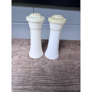 May include: Two vintage white salt and pepper shakers with light yellow lids. The shakers have a classic hourglass shape and are made of a smooth, opaque material. They are displayed on a wooden surface.