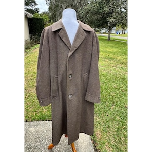 May include: A long, brown, textured overcoat featuring a classic collar, long sleeves with buttoned cuffs, and a front button closure. The coat is displayed on a mannequin, showcasing a structured silhouette with two side pockets and a subtle herringbone pattern throughout the fabric.