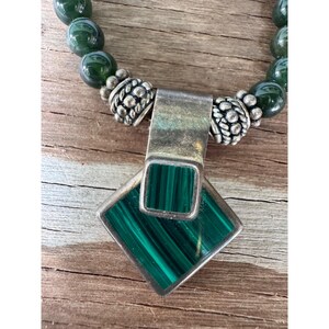 May include: A silver necklace with a malachite pendant and green beaded chain. The pendant is a square shape with a rectangular malachite stone. The necklace is on a wooden surface.