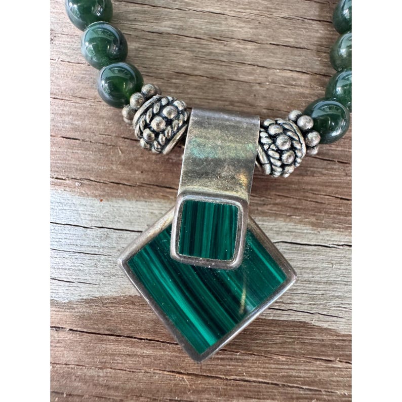 May include: A silver necklace with a malachite pendant and green beaded chain. The pendant is a square shape with a rectangular malachite stone. The necklace is on a wooden surface.
