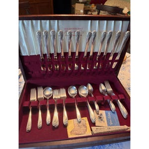 May include: A 1847 Rogers Bros. silver-plated flatware set housed in a dark wooden case with a burgundy velvet interior. The set includes knives, forks, and spoons with floral handles, along with informational cards and a small cleaning cloth.