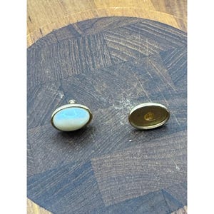 May include: A pair of oval-shaped, gold-tone cufflinks. One cufflink has a white, iridescent center, while the other cufflink has a gold center.