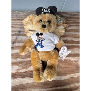 May include: A plush teddy bear with a black and white Mickey Mouse hat and a white t-shirt featuring Mickey Mouse in a blue and yellow outfit. The bear is light brown with a black nose and eyes. A heart-shaped tag is attached.
