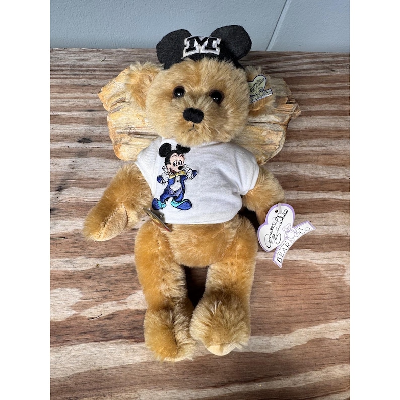 May include: A plush teddy bear with a black and white Mickey Mouse hat and a white t-shirt featuring Mickey Mouse in a blue and yellow outfit. The bear is light brown with a black nose and eyes. A heart-shaped tag is attached.