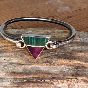 May include: Silver-toned bangle bracelet with a triangular pendant. The pendant features a green and purple stone inlay. The bracelet is set against a rustic wooden background.