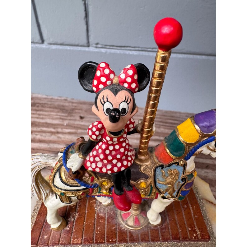 K&ouml;nnte beinhalten: Eine Minnie Maus Karussellfigur. Minnie tr&auml;gt ein rotes Kleid mit wei&szlig;en Punkten und eine passende Schleife. Das Karussellpferd ist wei&szlig; mit bunten Akzenten und einer goldenen Stange mit einer roten Kugel.