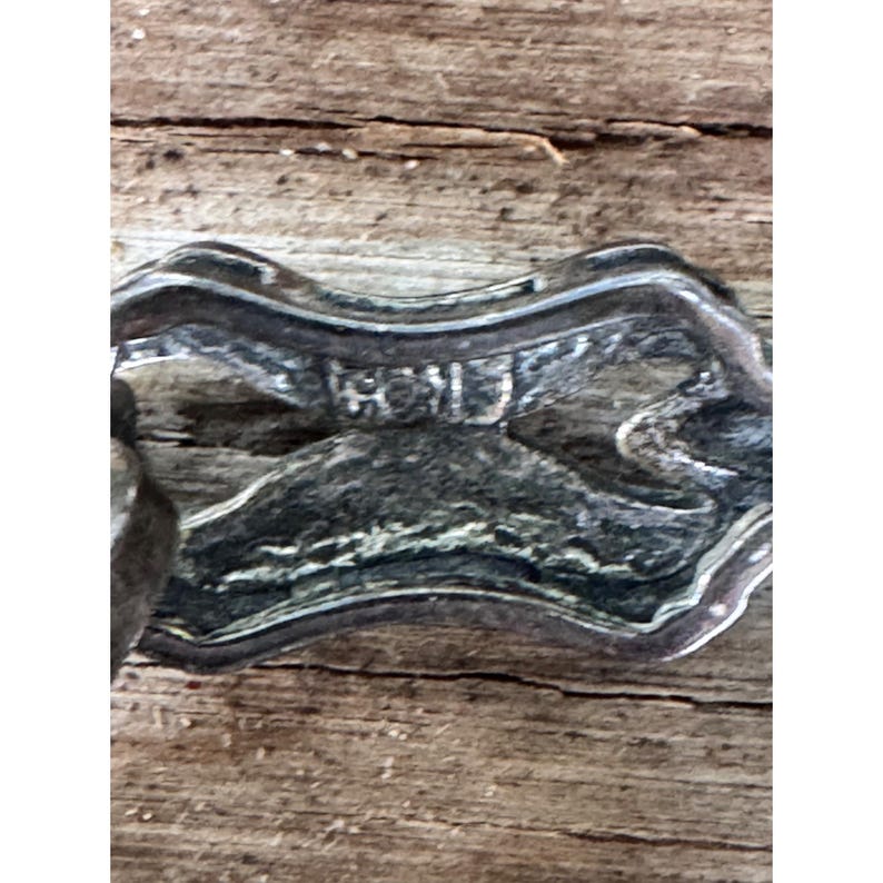 May include: Close-up of a silver-toned metal clasp with the word "HOLD" stamped on it. The clasp has a decorative, curved design and appears to be vintage. The background is a weathered wooden surface.