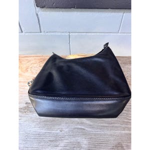 May include: A black leather handbag with a silver-toned chain detail. The bag has a trapezoid shape with a rounded top and a contrasting bottom panel. The bag is sitting on a wooden surface.