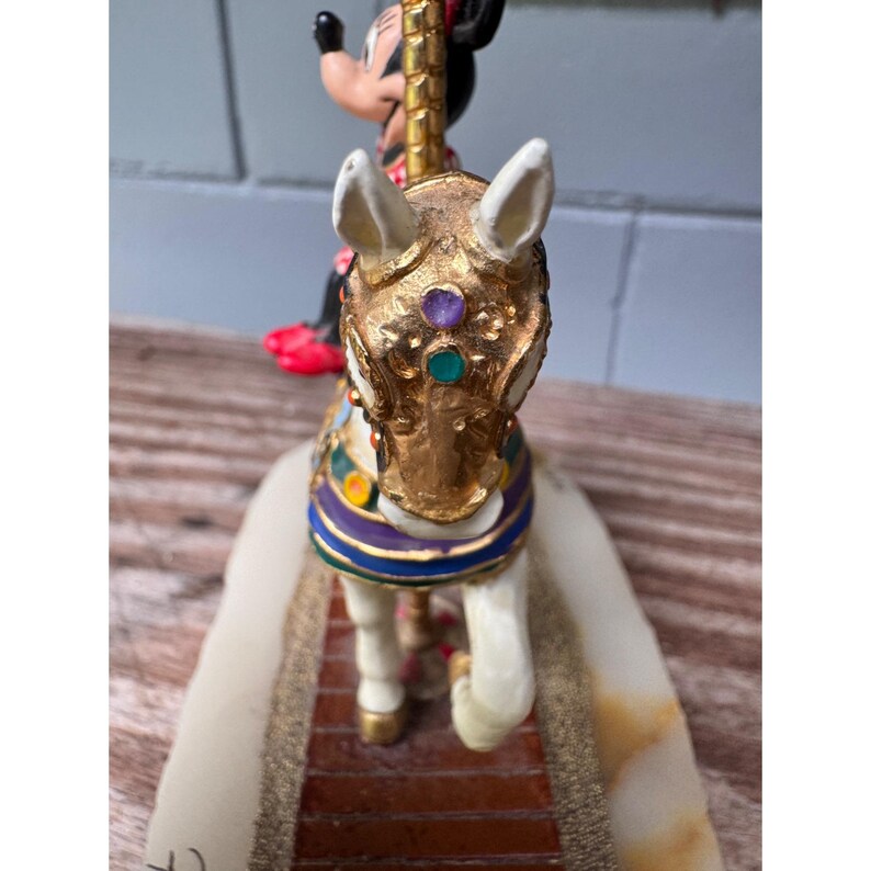 K&ouml;nnte beinhalten: Dekorative Figur eines wei&szlig;en Pferdes mit goldenem Kopf und aufwendigen Details. Eine Minnie-Maus-Figur reitet auf dem Pferd. Das Pferd hat einen blauen, lila und goldenen Sattel. Die Figur steht auf einem Sockel mit Ziegelmuster.