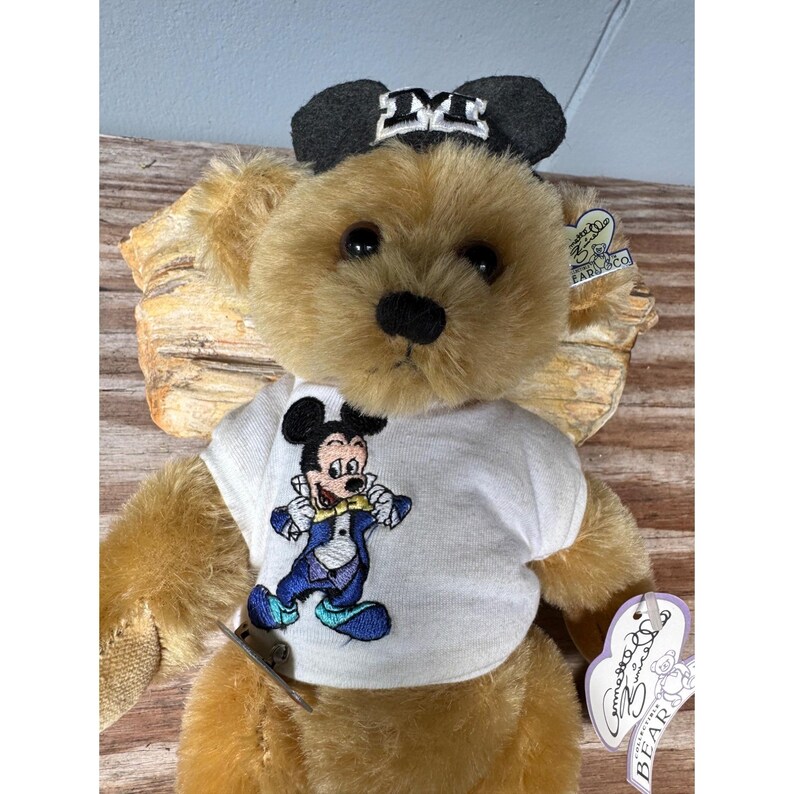 May include: A tan-colored teddy bear wearing a white t-shirt with a Mickey Mouse graphic and a black and white bow. The bear has a black nose and is holding a tag.