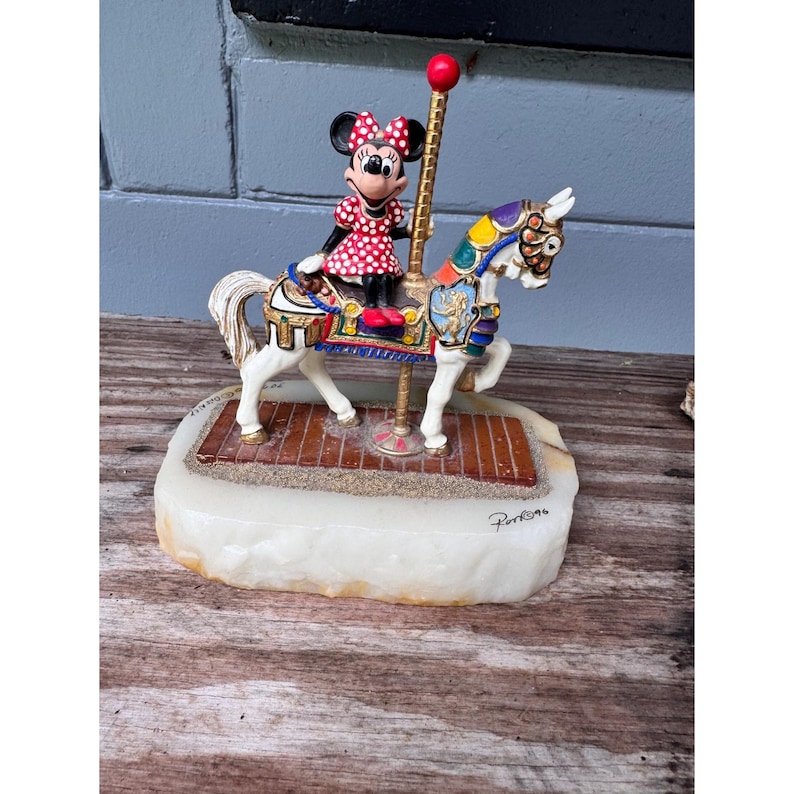 K&ouml;nnte beinhalten: Dekorative Karussellfigur mit Minnie Maus in einem roten gepunkteten Kleid, die ein wei&szlig;es Pferd mit bunten Akzenten reitet. Die Figur steht auf einem wei&szlig;en Sockel mit einer braunen Plattform und einer goldenen Stange.