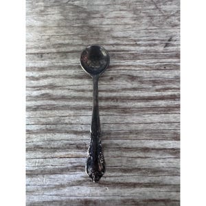 May include: A small, tarnished silver-toned metal spoon featuring an ornate, embossed floral pattern on the handle. The bowl of the spoon is round and shows signs of oxidation, while the handle tapers toward a decorative, detailed base.